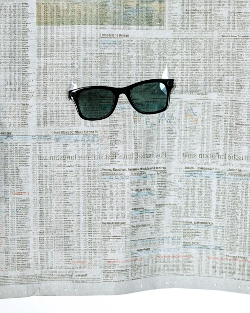 Black sunglasses taped to a crumpled newspaper sheet with financial data. The glasses are centered with tape visible at the top.