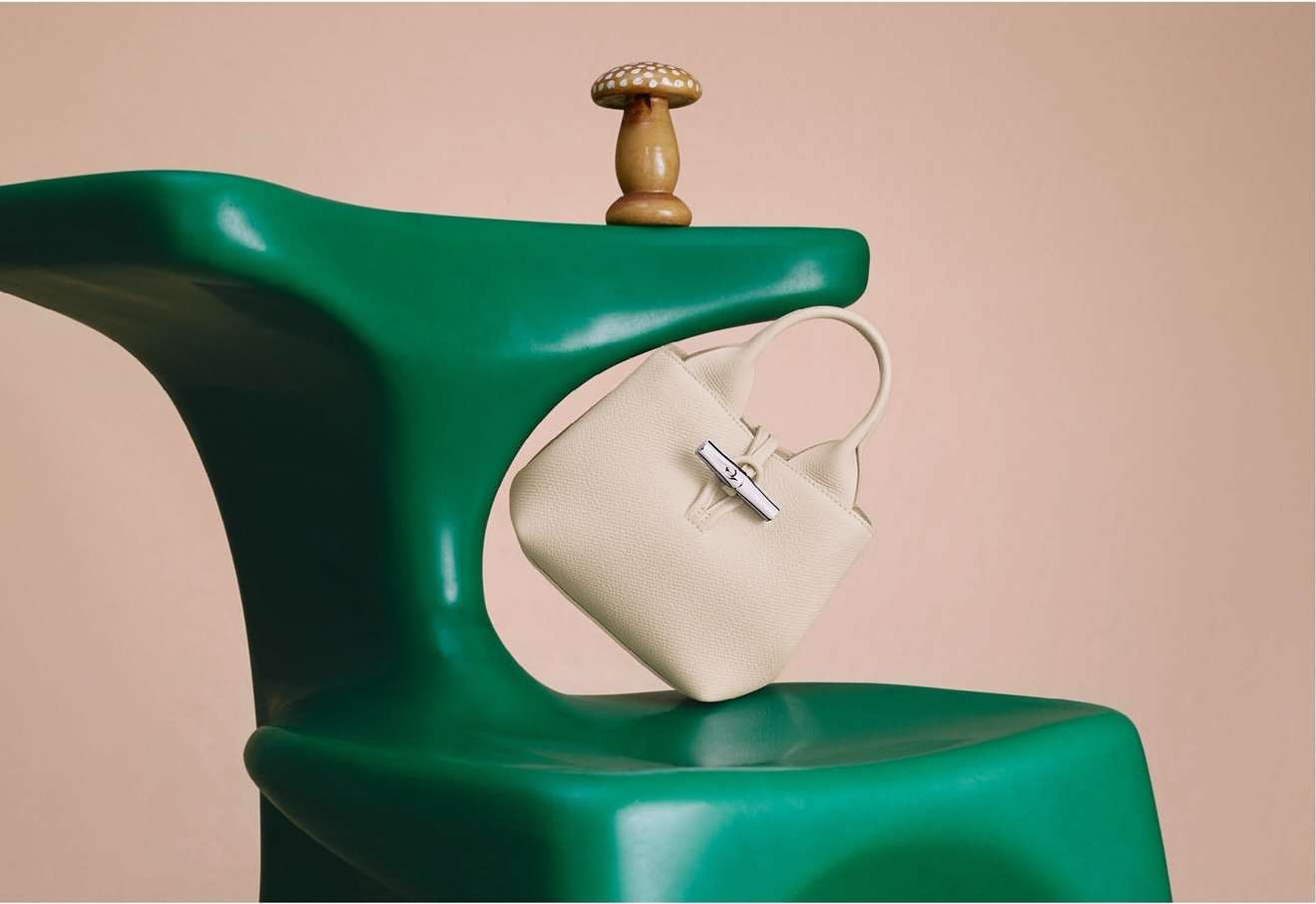 Ivory leather handbag on a green sculptural chair next to a wooden mushroom figurine, all against a plain peach background.