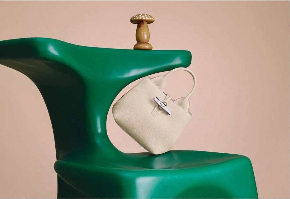 Ivory leather handbag on a green sculptural chair next to a wooden mushroom figurine, all against a plain peach background.