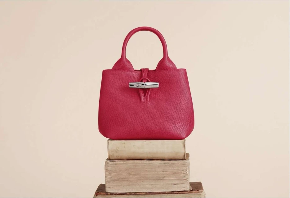 A red leather handbag with a silver clasp sits atop a stack of aged books against a pale-yellow background in a studio shot.