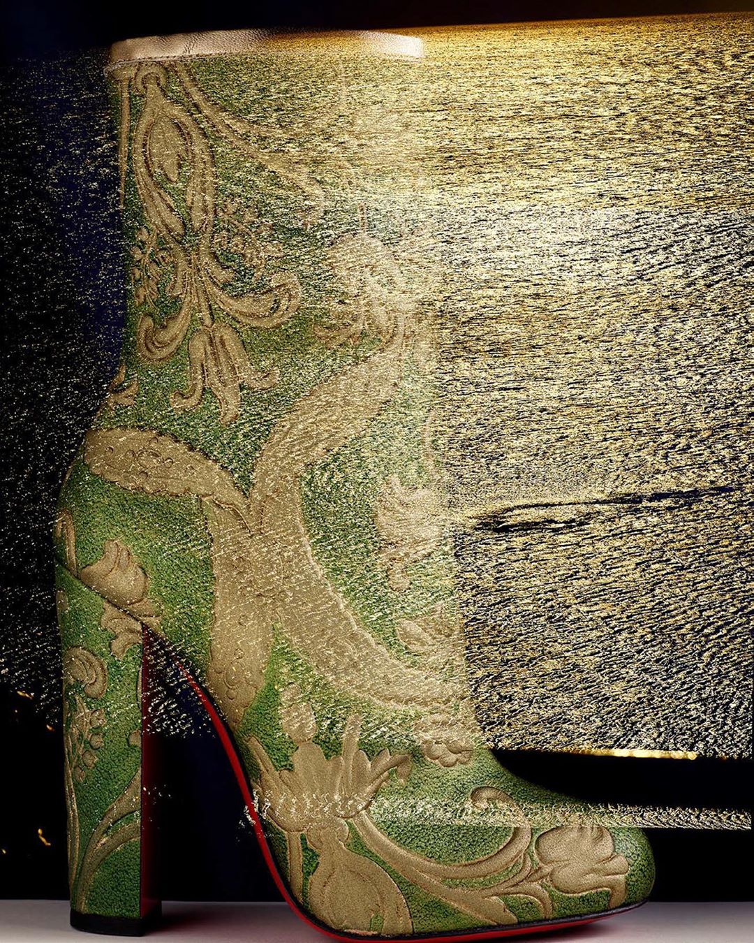 Embroidered high-heel ankle boot in shades of green and gold with red soles, against a blurred gold and black background.