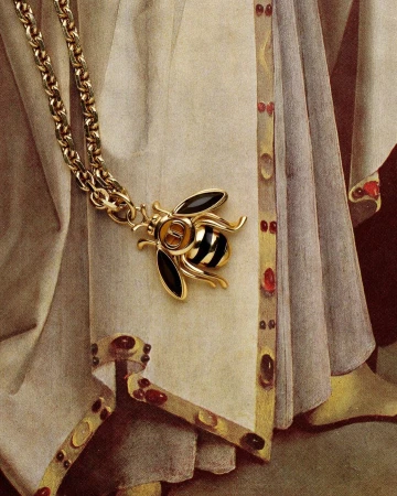 Close-up of a gold chain necklace featuring a bee pendant with black and gold stripes, draped over fabric with decorative trim.