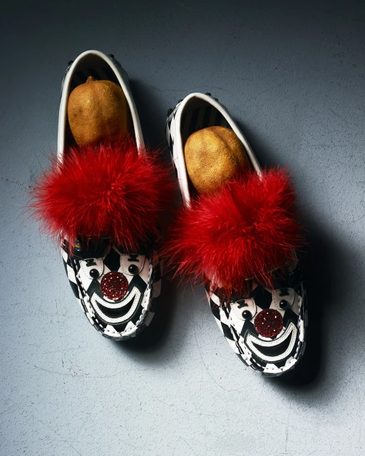Two clown-themed loafers filled with round objects, set against a neutral backdrop, creating a surreal and whimsical composition.