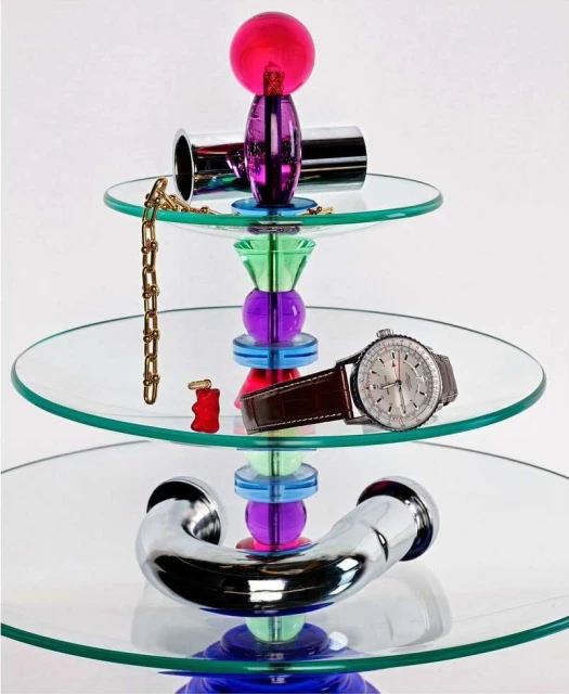 Three-tiered glass dessert stand with colorful plastic accents, featuring watch, gold chain, cufflinks, and gummy bear candy.
