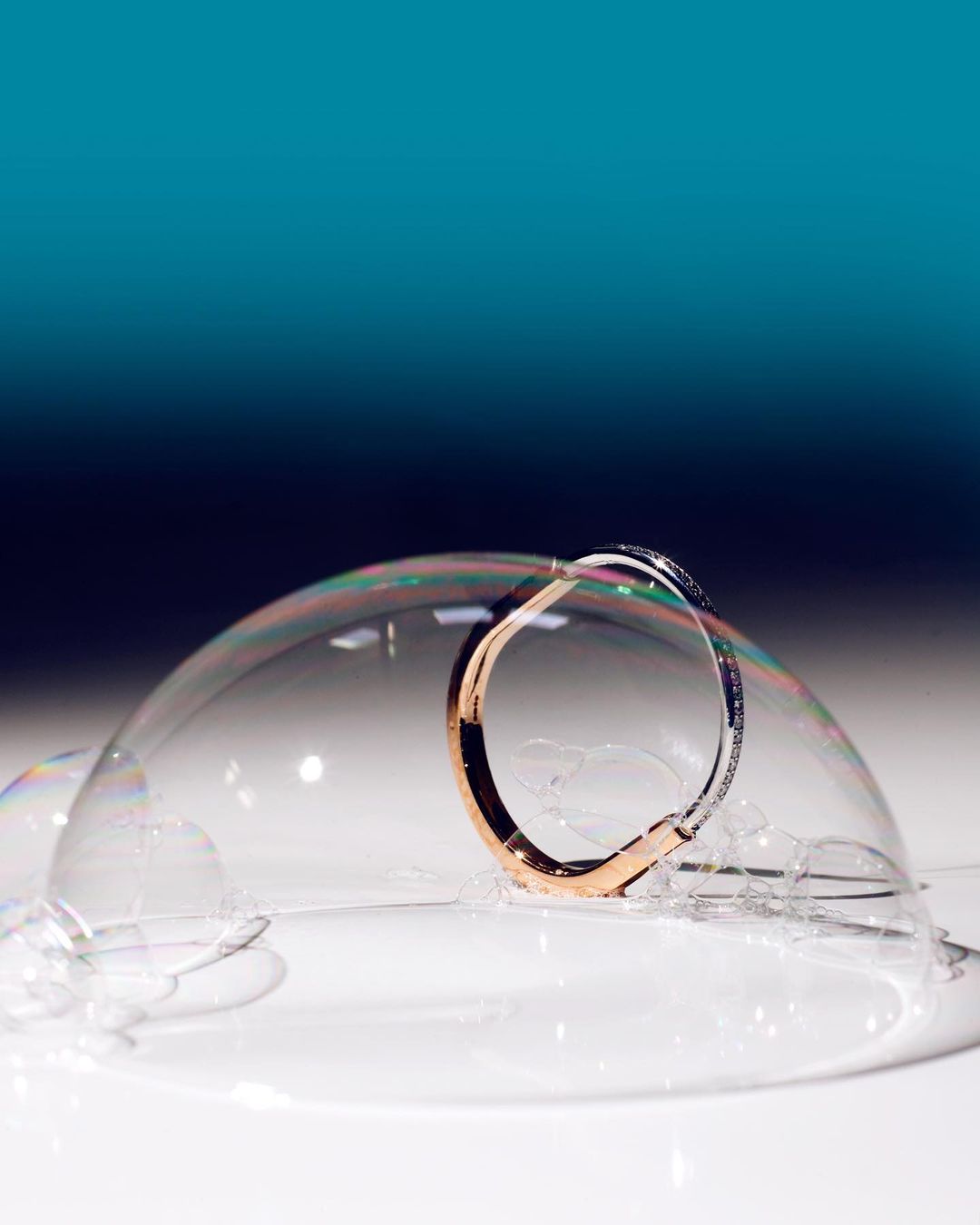 Close-up of a rose-gold and silver bangle encircled by a delicate soap bubble, set against a gradient teal background.