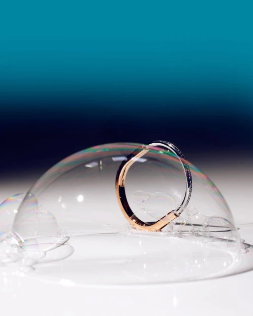 Close-up of a rose-gold and silver bangle encircled by a delicate soap bubble, set against a gradient teal background.