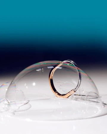 Close-up of a rose-gold and silver bangle encircled by a delicate soap bubble, set against a gradient teal background.