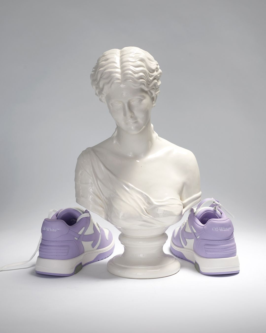 Still life shot of lavender and white sneakers flanking a Greco-Roman style bust on a neutral background. Fashion footwear.
