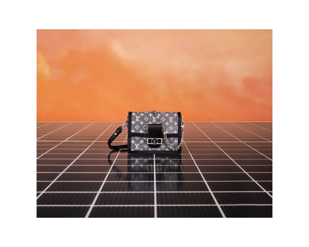 Monogram embossed leather handbag with chain strap in grey tones against an orange sky and black tile surface.