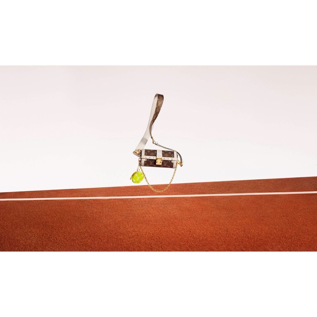 Luxury Louis Vuitton handbag featuring a lime-green tennis ball accessory, showcased against a vibrant red clay tennis court backdrop.