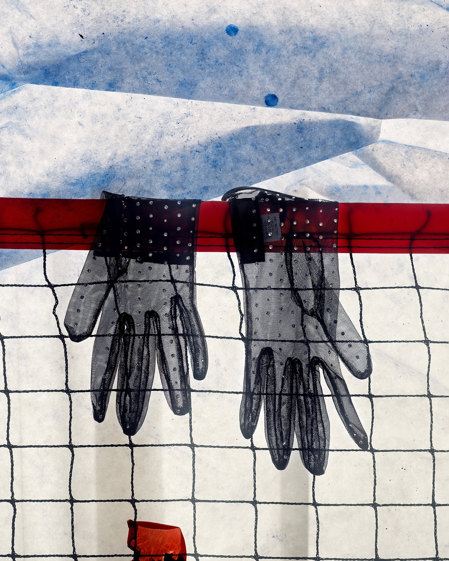 Pair of sheer black gloves with polka dots hanging on a red bar against a textured white and blue backdrop and black mesh screen.