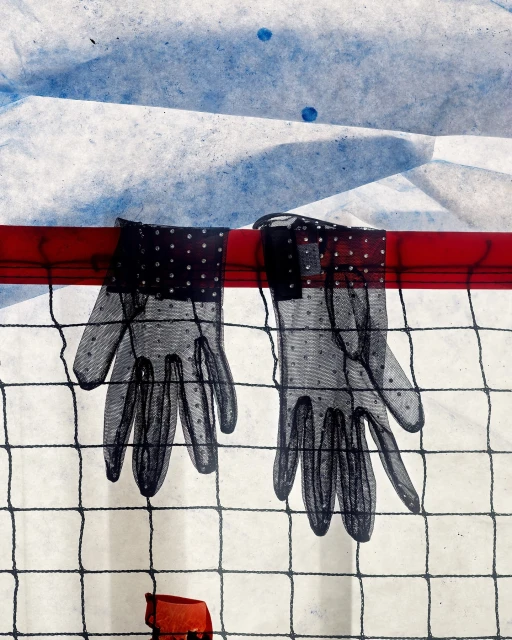 Pair of sheer black gloves with polka dots hanging on a red bar against a textured white and blue backdrop and black mesh screen.