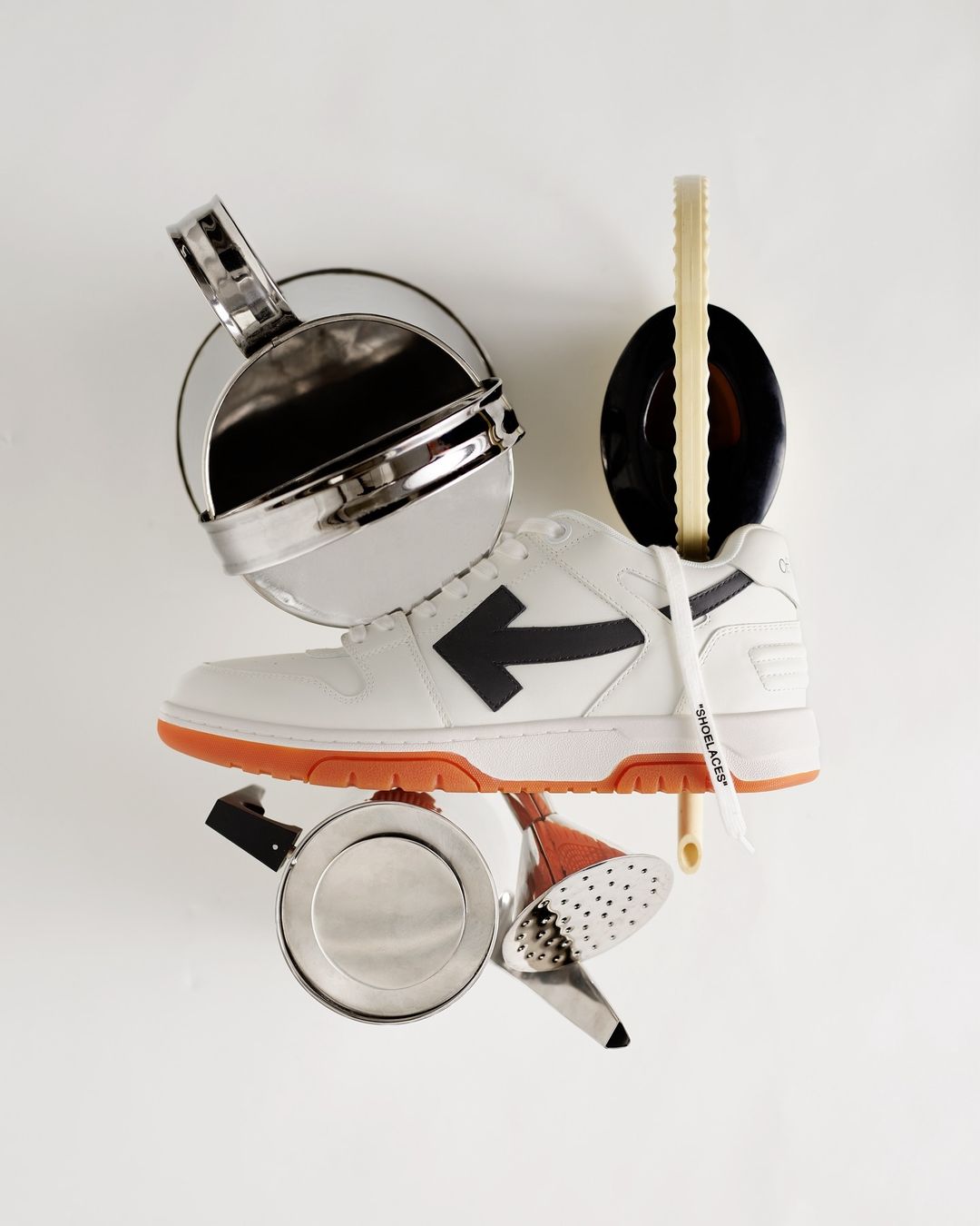 Studio shot of a white and orange Off-White sneaker surrounded by various silver kitchen objects against a white background.