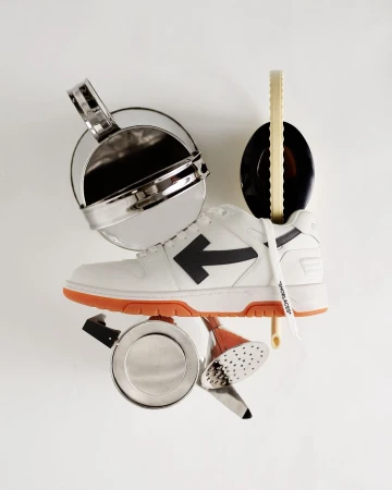 Studio shot of a white and orange Off-White sneaker surrounded by various silver kitchen objects against a white background.