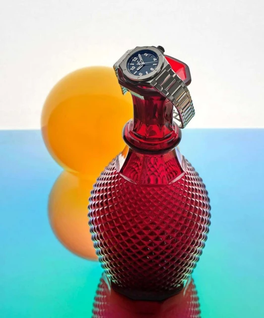 Silver wristwatch displayed on a red glass vase next to an orange sphere against a blue gradient background.