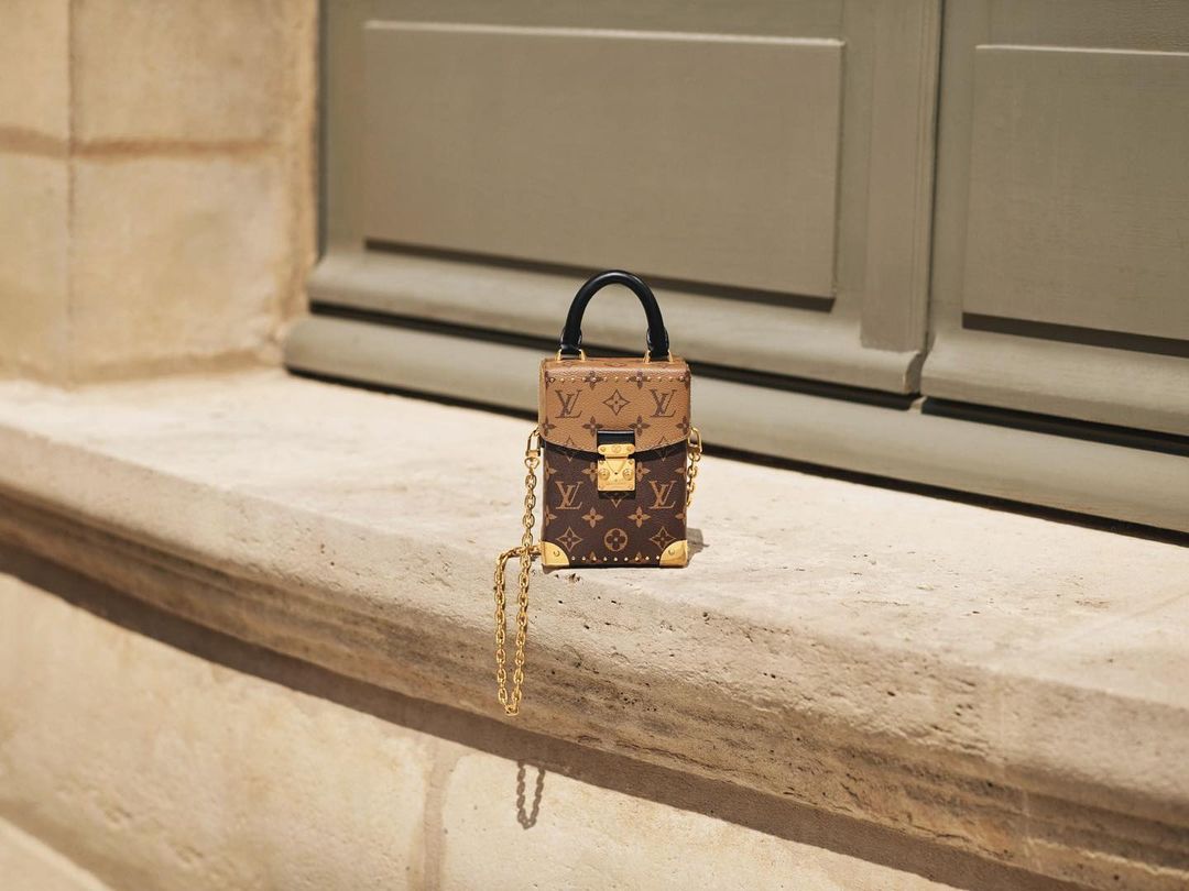 Close-up of a Louis Vuitton Petite Boite Chapeau handbag with gold chain on a stone ledge against a neutral wall.