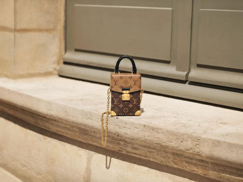 Close-up of a Louis Vuitton Petite Boite Chapeau handbag with gold chain on a stone ledge against a neutral wall.