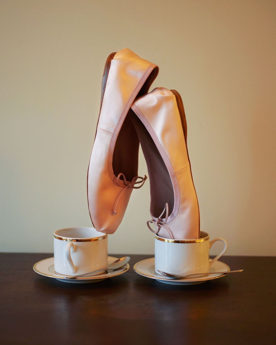 Still life of satin ballet shoes balanced inside of porcelain teacups with gold trim, set on a dark wooden surface in studio lighting.