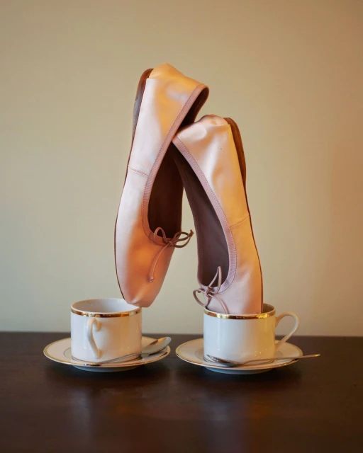Still life of satin ballet shoes balanced inside of porcelain teacups with gold trim, set on a dark wooden surface in studio lighting.