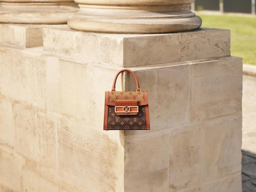 Brown monogrammed handbag with brown leather trim and gold hardware, set against a stone column and green foliage in the background.