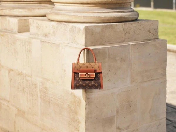 Brown monogrammed handbag with brown leather trim and gold hardware, set against a stone column and green foliage in the background.