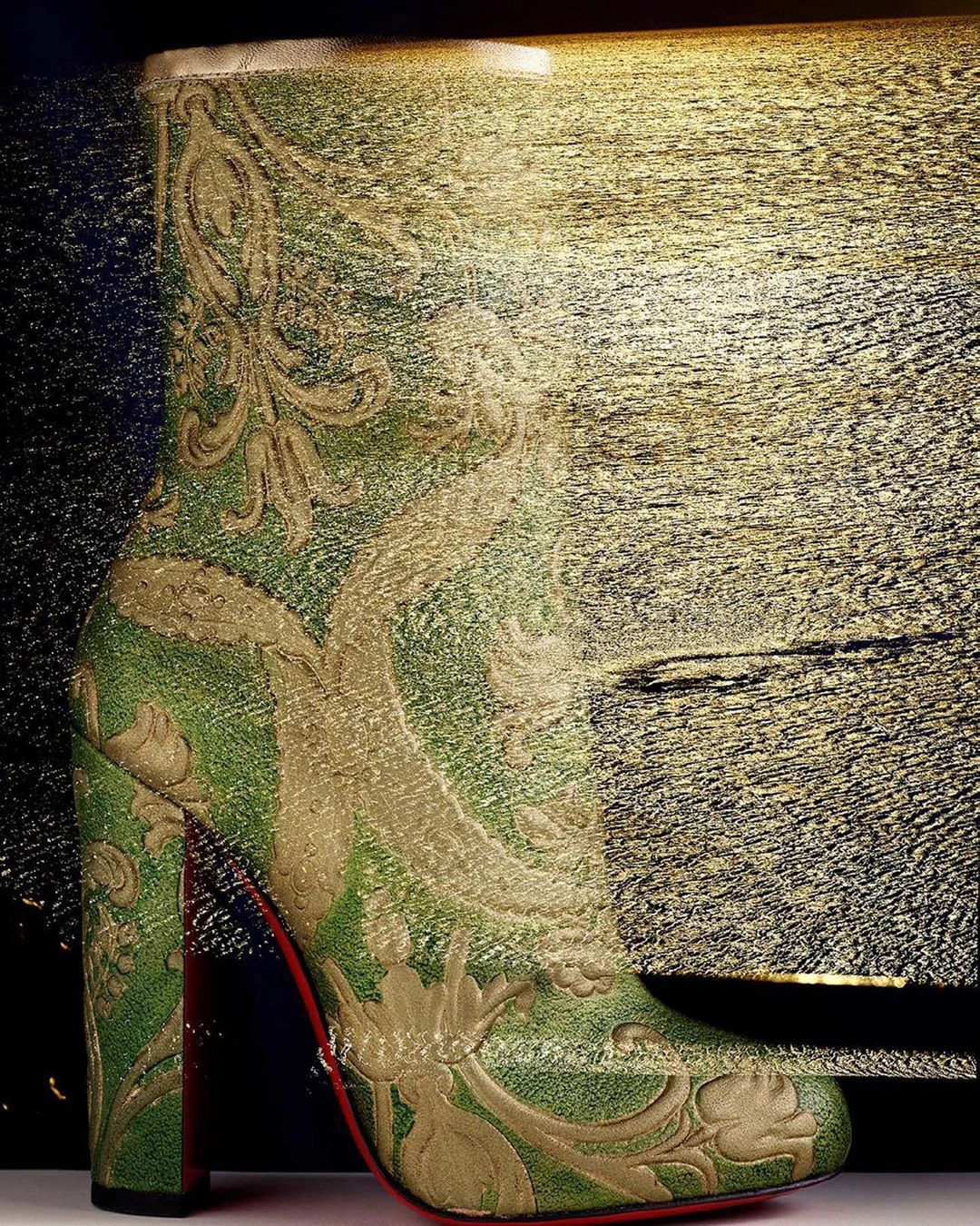 Close-up of an ornate green and gold brocade high-heeled boot with a signature red sole against a textured golden background.