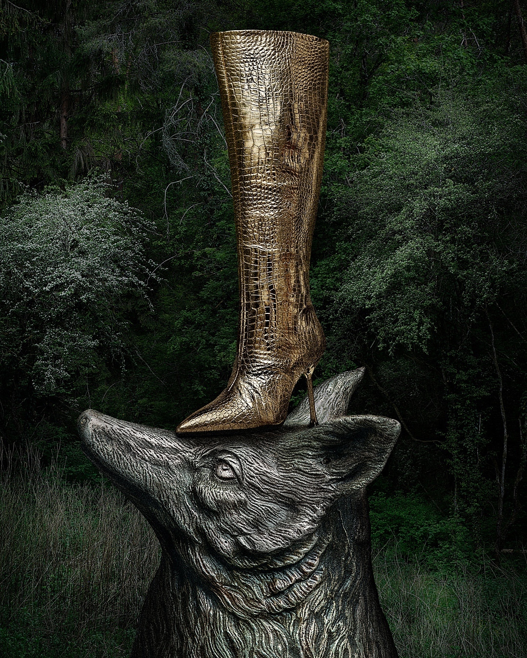 Gold crocodile-textured stiletto boot on top of a grey stone boar head statue with lush green foliage in the background.