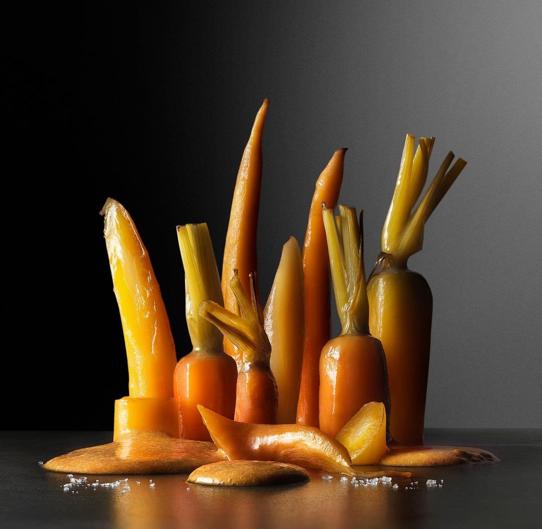Still life photograph of a group of glazed and peeled carrots arranged on a reflective surface with dark gradient background. Food photography.