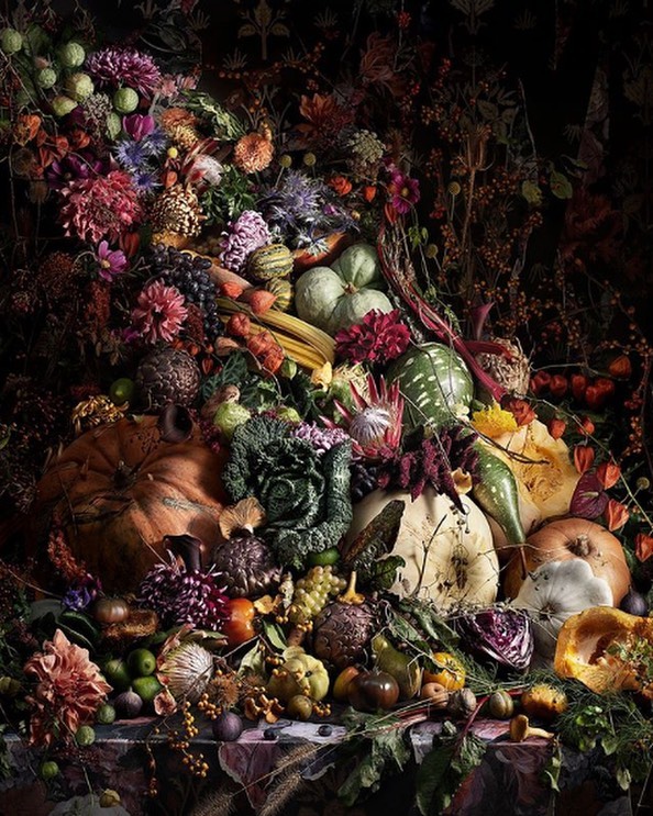 Elaborate still life with pumpkins, gourds, flowers, grapes, and autumn foliage on a dark surface with a backdrop of floral wallpaper.