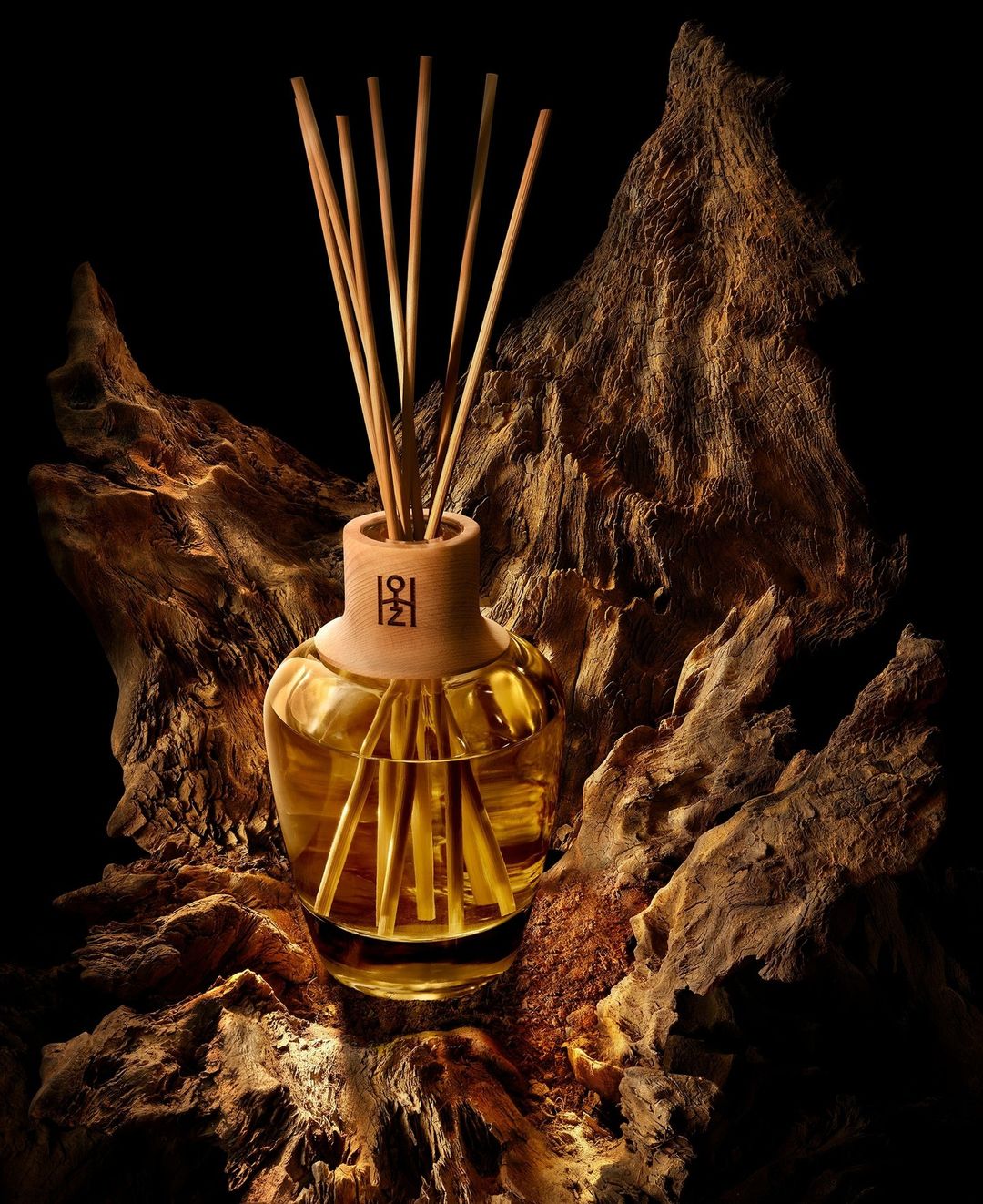 Reed diffuser with golden liquid in a glass bottle on top of a textured brown wood piece against a dark background.
