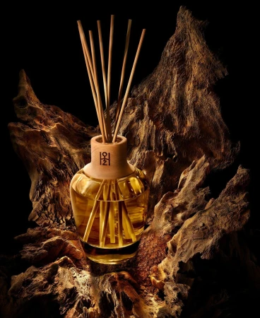 Reed diffuser with golden liquid in a glass bottle on top of a textured brown wood piece against a dark background.