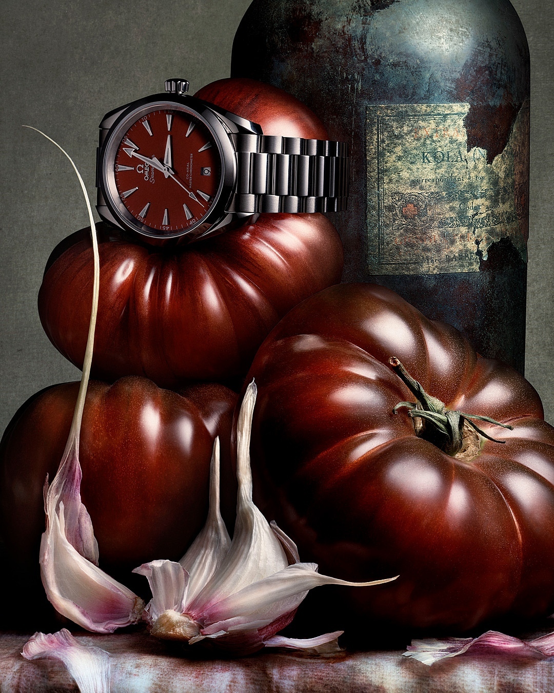 Still life shot of an Omega watch atop red tomatoes beside a dark bottle and garlic cloves in a moody, painterly indoor setting.