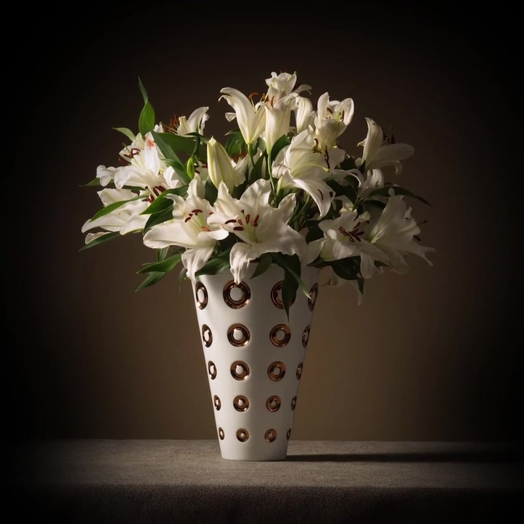 Still life photograph of white lilies in a white vase with gold accents against a dark background, on a dark surface.
