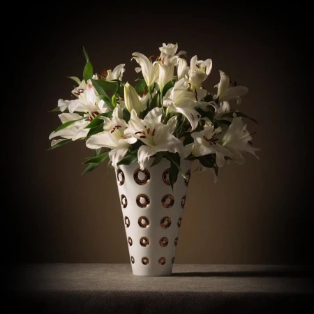 White lilies in a patterned white vase on a dark surface with dramatic lighting
