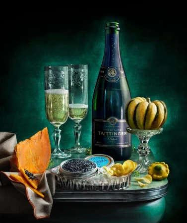 Still life photography featuring a bottle of champagne, two flutes, caviar, sliced squash, and potato chips against a dark green background.