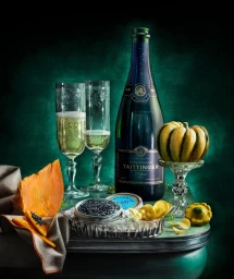 Still life photography featuring a bottle of champagne, two flutes, caviar, sliced squash, and potato chips against a dark green background.