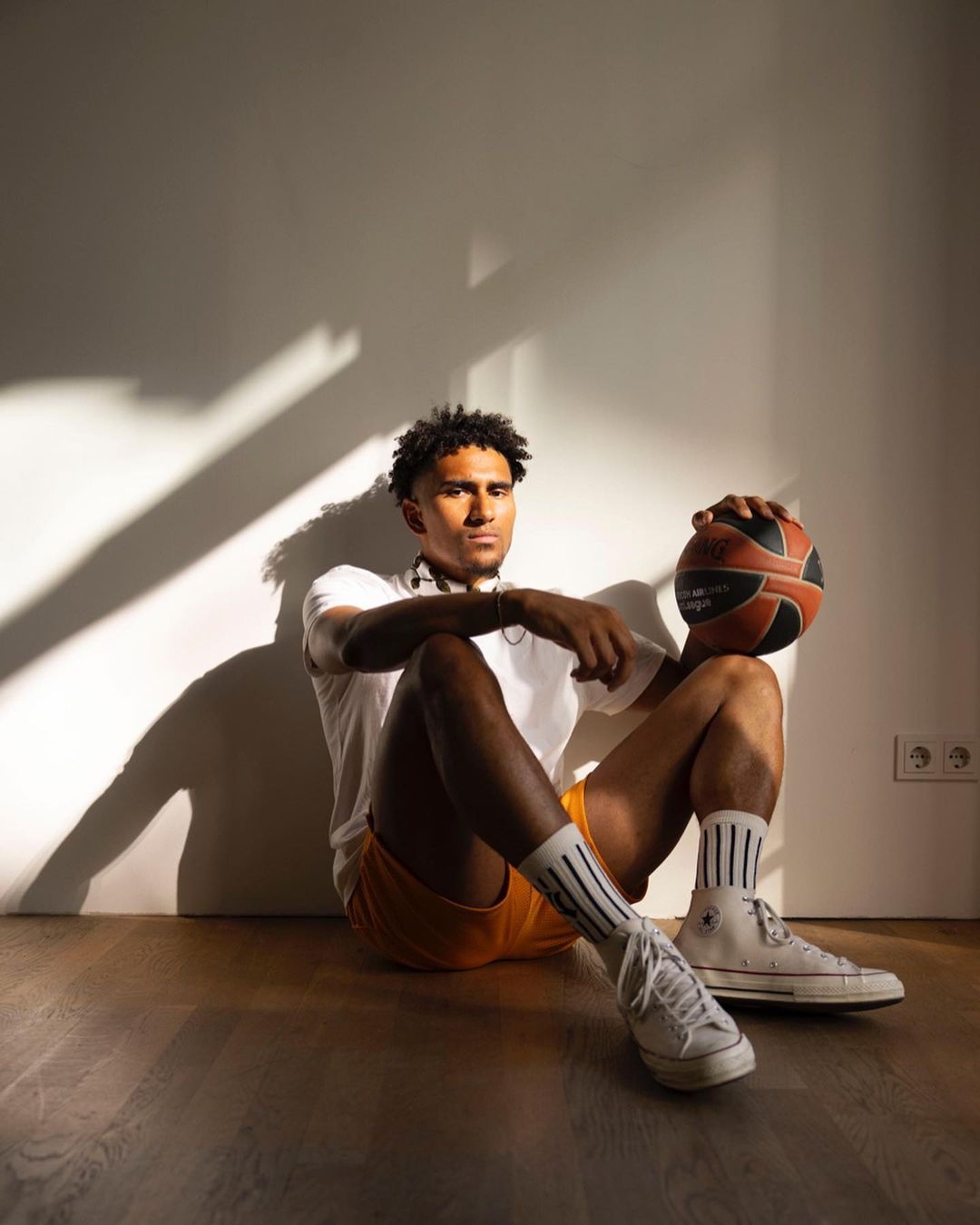 A young adult male with a basketball sitting indoors against a wall. He wears athletic socks, sneakers, and casual shorts and t-shirt.