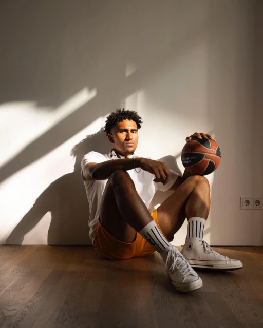 A young adult male with a basketball sitting indoors against a wall. He wears athletic socks, sneakers, and casual shorts and t-shirt.