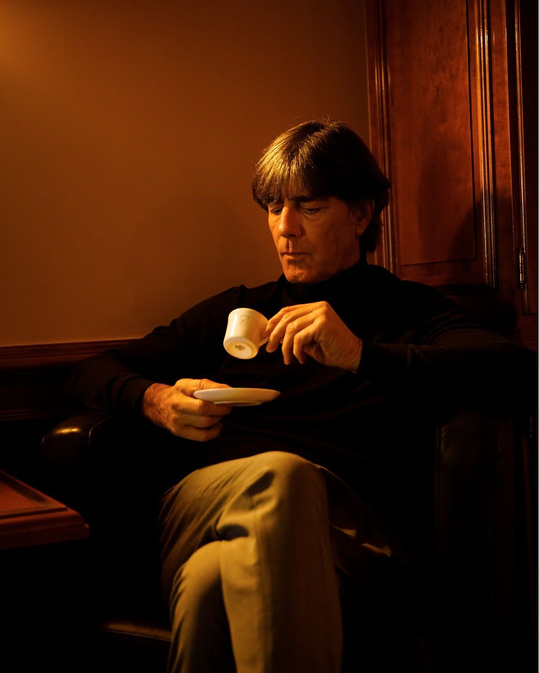 An adult man with dark hair sits in a dimly lit room, holding a white tea cup and saucer. He is wearing a black turtleneck sweater.