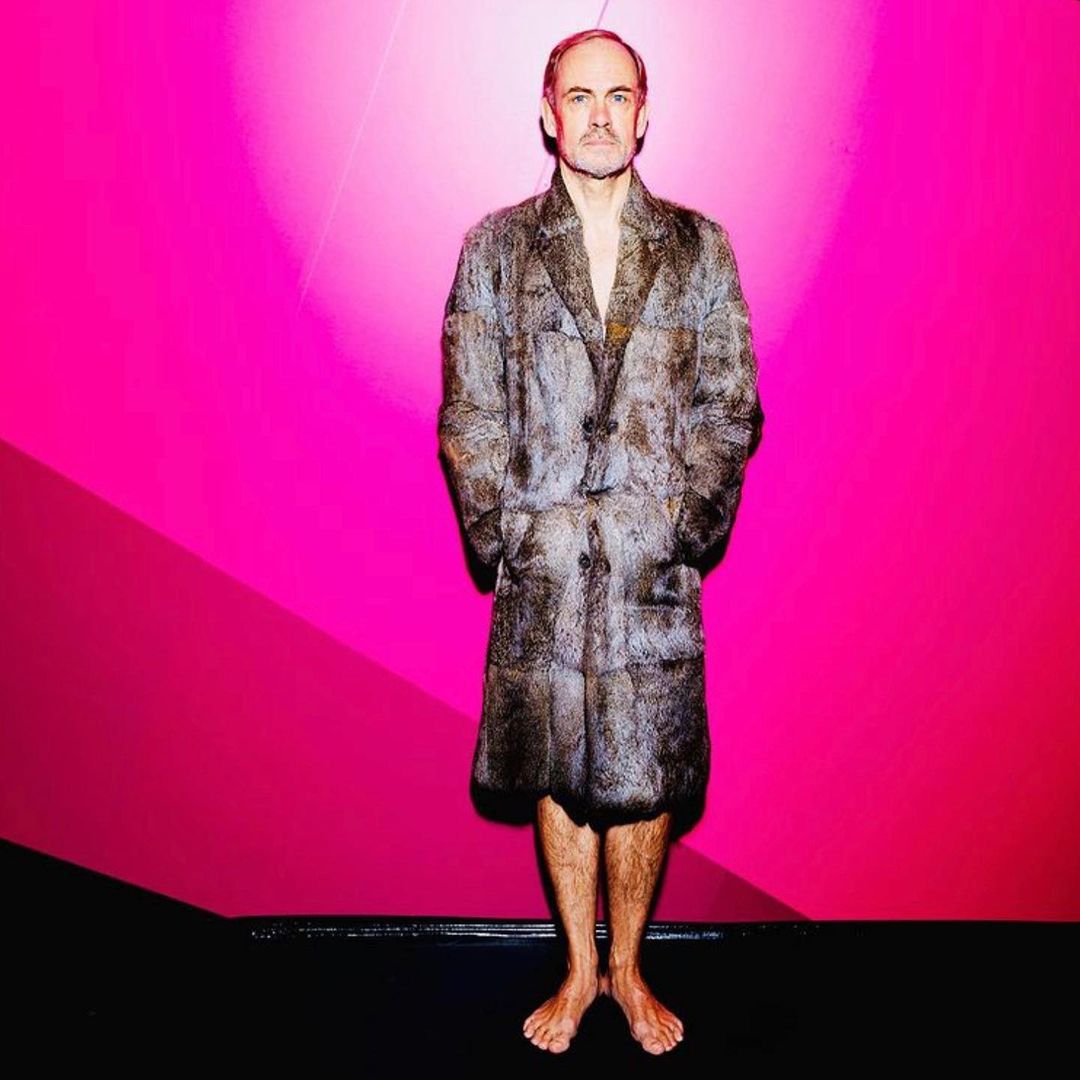 Full-length portrait of a man in a fur coat, barefoot, standing in front of a pink wall with a black floor for fashion editorial.