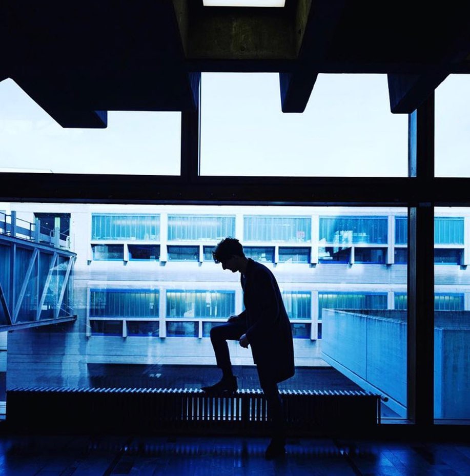 Silhouette of a slender adult male standing on a metal radiator by a window, looking out onto an urban landscape and architecture.