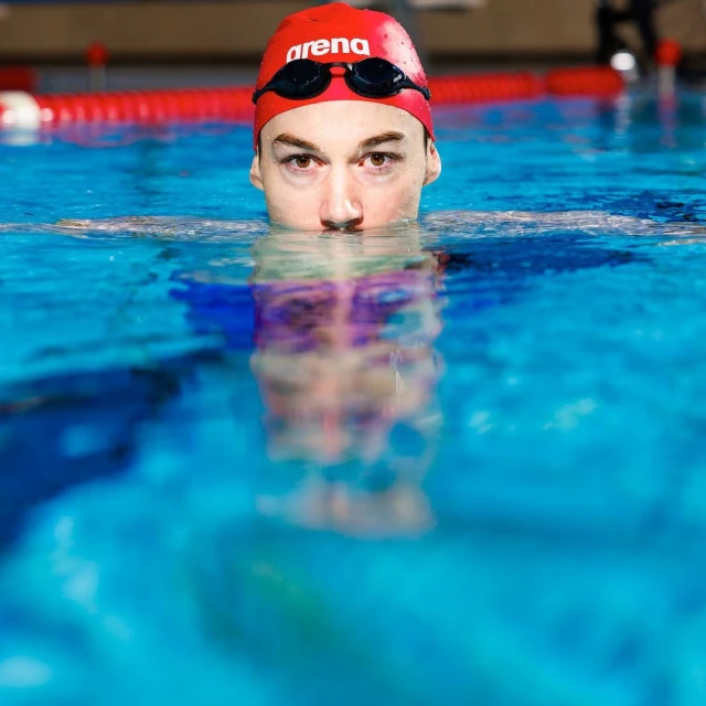 Close-up of a swimmer wearing a red cap and goggles in a pool. Focus on the swimmer's face and upper body above the water.