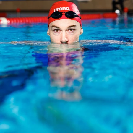 Close-up of a swimmer wearing a red cap and goggles in a pool. Focus on the swimmer's face and upper body above the water.