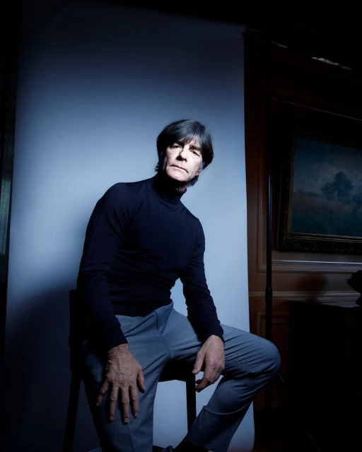 Portrait of a mature man seated on a chair in a studio. He is wearing a black turtleneck and gray pants.  Dark interior with painting.