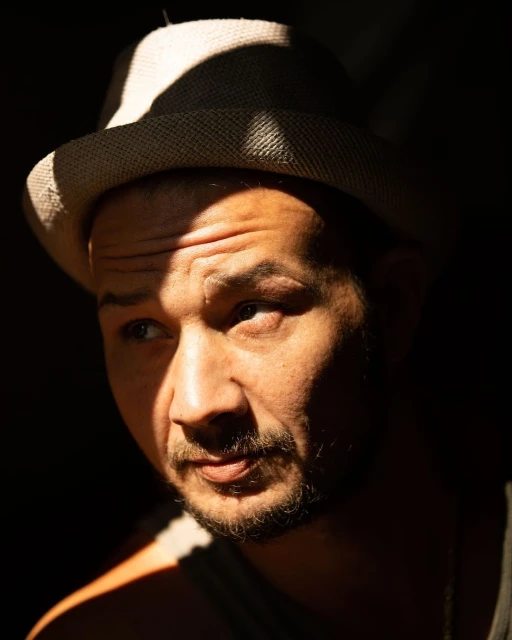 Portrait of a man in shadows wearing a hat, with light emphasizing his face and upper body against a dark background.
