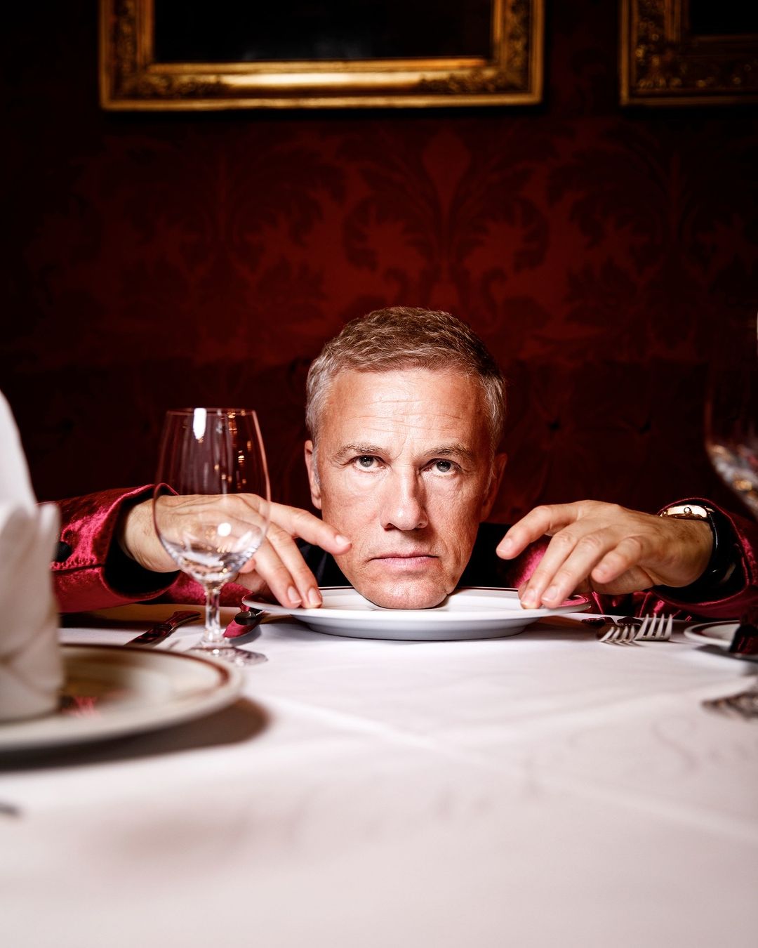 Headshot of adult male at the head of a dinner table, wearing a red jacket and resting his chin on a white dinner plate.