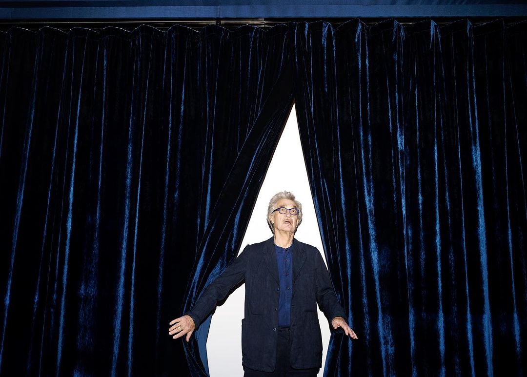 Elegant mature man with glasses stands in front of dark blue velvet curtains, wearing a navy jacket and blue shirt, indoors.