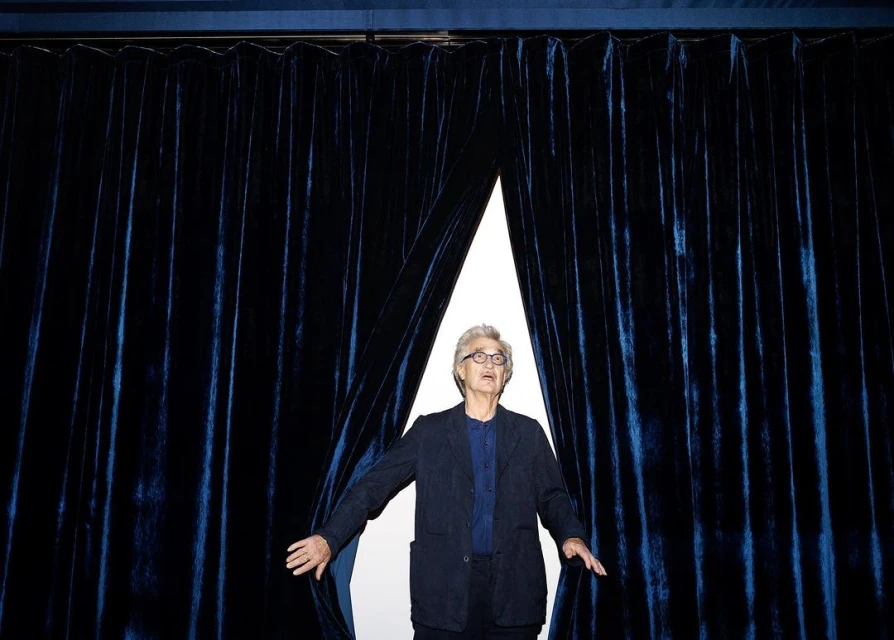Elegant mature man with glasses stands in front of dark blue velvet curtains, wearing a navy jacket and blue shirt, indoors.