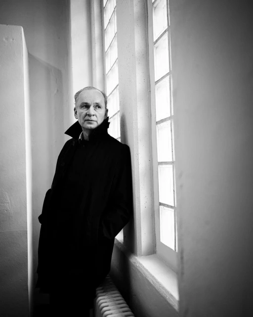 Black and white portrait of a man in a dark coat, standing indoors beside a bright window with multiple panes.