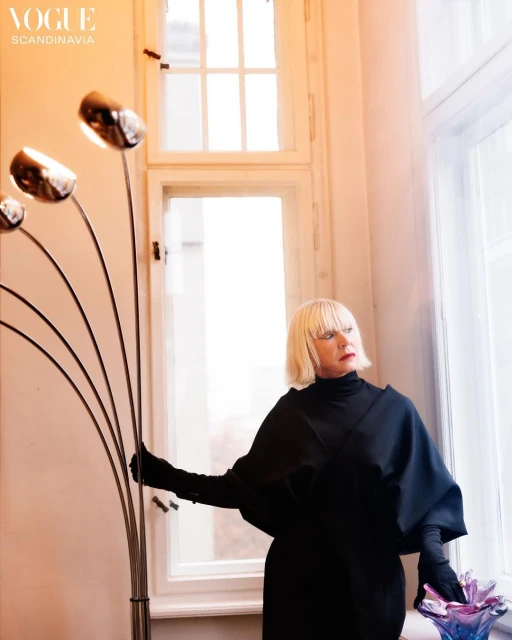 Mature woman with blonde hair poses in black dress and gloves by window and unique lamp in soft light, high fashion editorial.